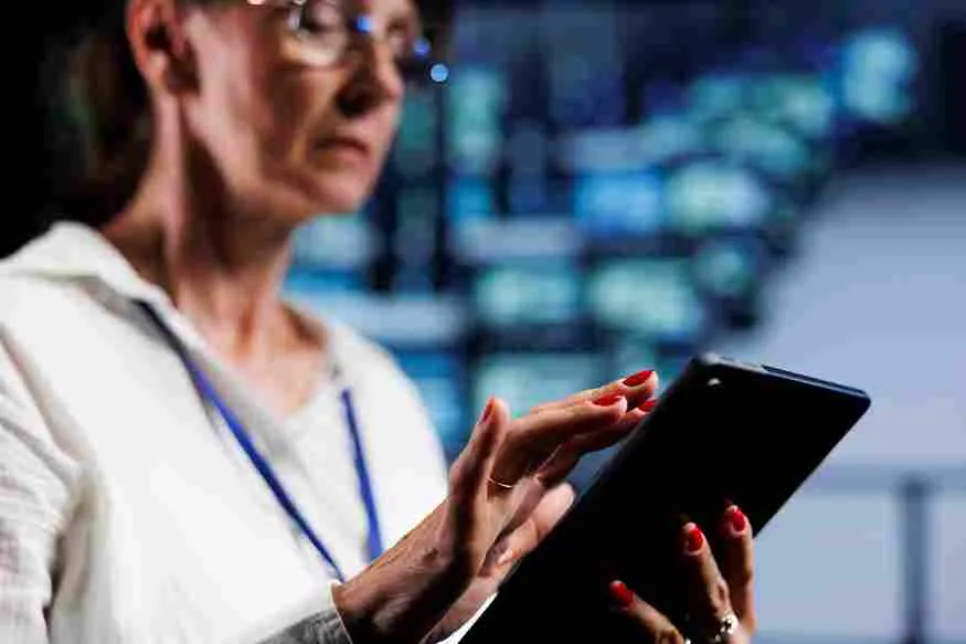 Mulher com óculos e traje profissional opera um tablet em um ambiente de alta tecnologia com múltiplos monitores ao fundo exibindo dashboards de segurança e vigilância. A imagem ilustra o uso de tecnologia antiperdas e inteligência artificial no varejo alimentar para monitoramento de inventário, prevenção de furtos e controle de quebras operacionais em tempo real.