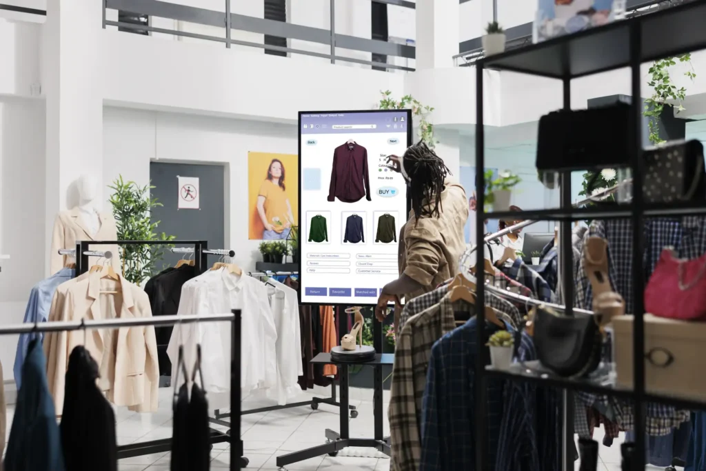 Homem com dreadlocks interage com um totem digital interativo em uma loja de roupas moderna. A tela exibe um catálogo de camisas masculinas com opções de cores e botão de compra. Ao redor, araras com roupas e prateleiras de acessórios em um ambiente de loja física conceito.