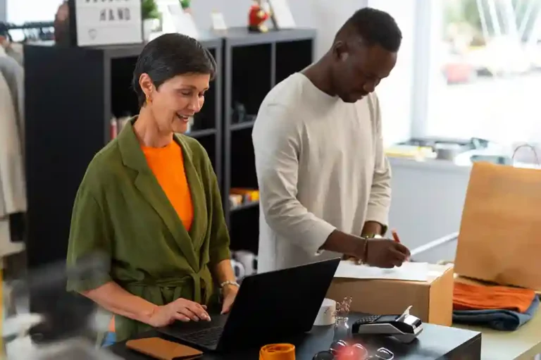 Visão de um ambiente de loja física com dois colaboradores realizando tarefas operacionais. À esquerda, uma mulher de blusa laranja e casaco verde sorri ao operar um notebook em um balcão. À direita, um homem de blusa bege prepara um pacote para envio, escrevendo em uma etiqueta. Sobre a mesa, há uma máquina de cartão e embalagens. A imagem ilustra o funcionamento do backoffice para e-commerce, demonstrando a integração necessária entre a gestão de pedidos, estoque e expedição para garantir a eficiência operacional e o sucesso das vendas online.