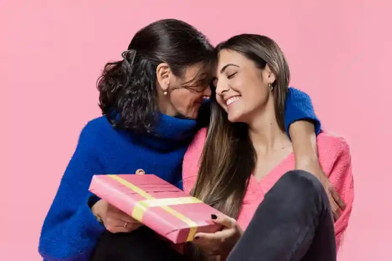 Mãe e filha em um momento afetuoso, sorrindo e abraçadas contra um fundo rosa. A filha, vestindo um suéter rosa, entrega um presente embrulhado para a mãe, que usa um suéter azul vibrante. A imagem ilustra a celebração e a troca de presentes no Dia das Mães, servindo como conceito visual para campanhas de marketing de e-commerce e estratégias de vendas sazonais em 2026