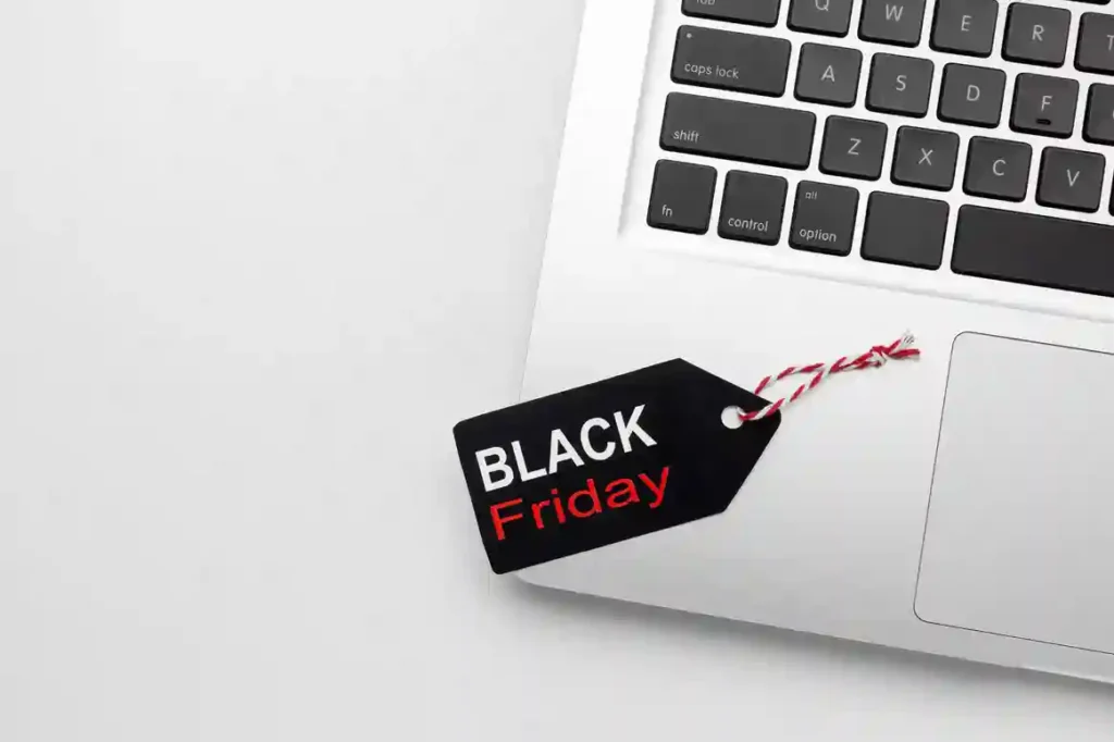 Visão superior de parte de um notebook prateado com teclado preto sobre uma mesa branca. Ao lado do teclado e do trackpad, repousa uma etiqueta de preço preta amarrada com um cordão listrado vermelho e branco, onde se lê a inscrição 'BLACK FRIDAY' em letras brancas e vermelhas. A imagem ilustra a preparação de e-commerces e lojas virtuais para a Black Friday 2026, destacando campanhas promocionais, planejamento de estoque e estratégias de vendas digitais.